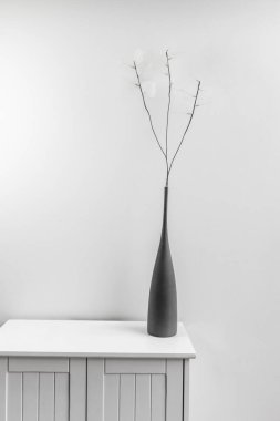 İnce beyaz yapraklı yapay dalları olan gri vazosu olan küçük beyaz ahşap bir dolap.