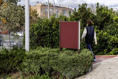 Uzun saçlı bir adam çalılar ve ağaçlarla dolu bir şehir parkında yürüyor.