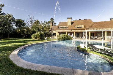 Ortasında bir çift Balili yatak ve dekoratif dikey su jetleri olan büyük bir havuz. Arka planda büyük bir aile evi var.