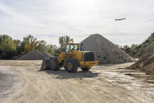A large piece of construction machinery next to large mounds of river sand and a plane flying over the scene