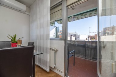 Kahverengi teraslı küçük bir teras, yeşil boyalı metal korkuluklar, diğer binaların manzarası ve cam yemek masası olan oturma odasına giriş.