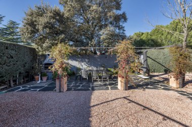 Çalılar ve bitkiler arasında tabanı kumla kaplı tek kişilik bir ev ve yemek masası..