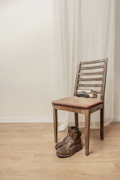 Brown leather mountaineering boots resting under the seat of a emaciated wooden chair next to an old camera with some books and a subtle white curtain behind it
