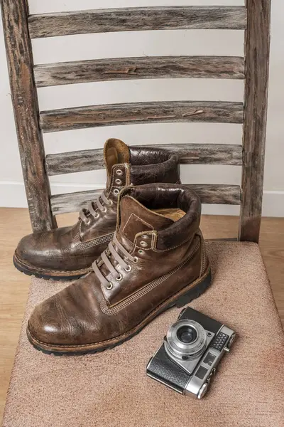 A pair of men\'s brown leather boots with triple stitching on an old chair next to a vintage photo camera in a room with brown parquet floors and white walls