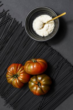 Lezzetli, olgun Raf domates üçlüsü bir yığın siyah makarna üzerinde pürüzsüz, siyah bir yüzey üzerinde rendelenmiş peynirle dolu bir kasenin yanında.