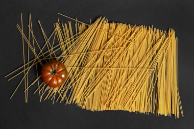 Pürüzsüz siyah bir yüzeye yayılmış büyük bir buğday spagettisi yığını üzerinde parlak raf domatesi.