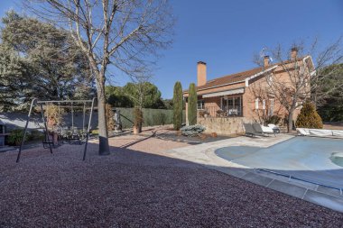 Çakıl taşı zeminli müstakil dağ evi tipi arsa, içinde selvi ağaçları, mavi tuval ve gri metal çocuk salıncaklarıyla kaplı yüzme havuzu.