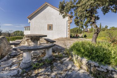 Beyaz cepheli bir kır evi, arsada taş döşemeler ve çimento bankları olan bir masa.