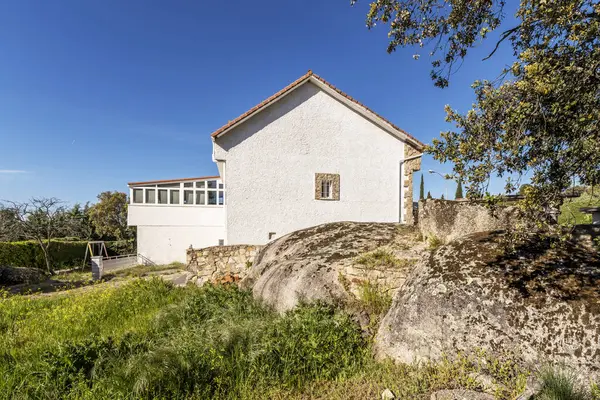 A multi-story country house with white concrete facades, large windows and terracotta roofs, large granite boulders on the floor and clear blue skies