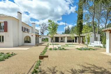 Balkonlarda kırmızı ahşap panjurları olan beyaz bir kır evinin ön cephesi ve bulutların içinden geçen parlak bir günde çakıllı zemini olan yeni dikilmiş bahçeler.