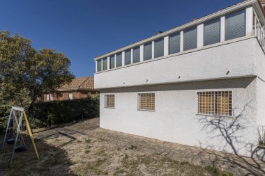 Beyaz duvarları ve taş zemini olan bir dağ evinin arka cephesi, çitlerle çevrili ve koltuk olarak tekerlekleri olan çocuk salıncakları.