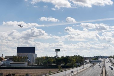 Bulutlu gökyüzünde yükselen bir uçak. Bir otoyol ve bira fabrikasının yanında.
