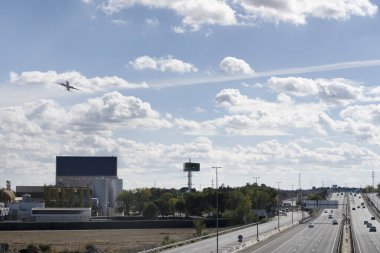 Bulutlu gökyüzünde yükselen bir uçak. Bir otoyol ve bira fabrikasının yanında.