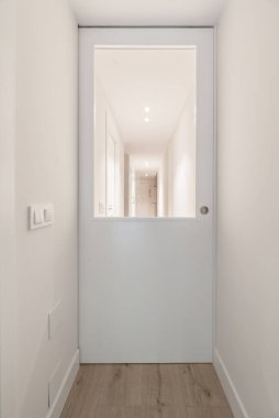 Sade beyaz duvarları olan bir evin koridoru, cam pencereli beyaz lakaplı kaplar ve aynı renkte ışık düğmeleri ve açık ahşap döşemeler.