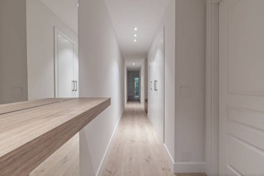 Salonu pürüzsüz, beyaz duvarları olan, koridoru cam pencereli beyaz, cam kapılı bir dairede açık tahta büfe ve ayna.