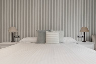 Yatak odasındaki komodinlerin yanında ikiz lambalar, çeşitli yastıklar ve dekoratif kağıtlarla kaplı bir duvar.