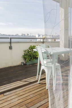 Ahşap zeminleri, yeşil bitkileri ve şeffaf perdeleri olan şehir terasında uyumlu sandalyeleri olan güzel bir pastel mavi metal bahçe masası.