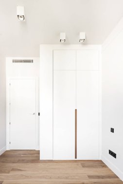 Boş, modern bir yatak odası. Beyaz kapıları ve ahşap renkli çivi çekmesi olan bir gardırobu var.
