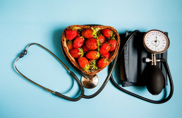 stock image The concept of health. Fresh strawberries with a stethoscope and a pressure measuring device on a blue background, top view