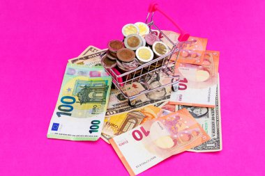 Bozuk paralarla dolu mini alışveriş sepeti ve pembe arka planda dolar banknotları. Finans ve tasarruf kavramı.