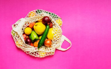 White string bag with different vegetables and fruits on a pink background. top view