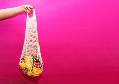 Kadınlar, pembe arka planda farklı sebzeler ve meyvelerle beyaz ip torbalarını tutarlar..