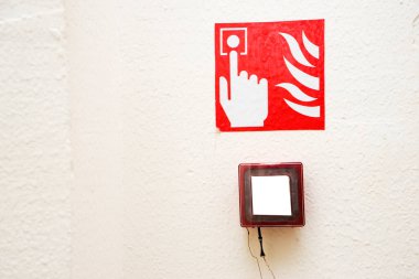 Fire alarm button encased in glass over a sign showing a finger pushing the button near flames on a white wall clipart