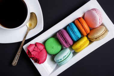 Beyaz bir fincan kahve ve siyah arka planda beyaz bir tabakta çok renkli badem kurabiyesi. Fransız gelenekleri