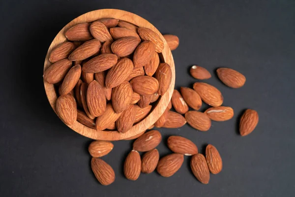 Peeled almonds in a wooden bowl and some scattered nuts on black background, top view.