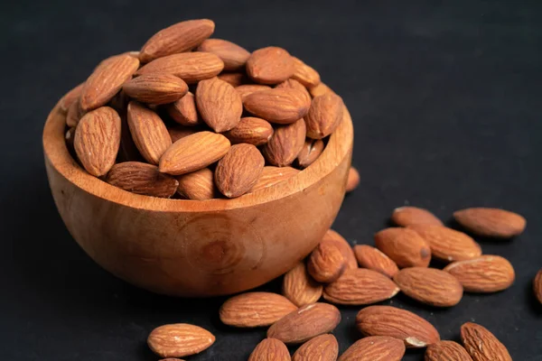 Peeled almonds in a wooden bowl and some scattered nuts on black background