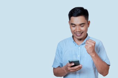 Genç kendine güvenen heyecanlı Asyalı genç adam açık mavi arka planda izole edilmiş akıllı telefona bakıyor. Kazanan konsept