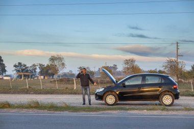 Genç Latin bir adam arabasının önünde cep telefonuyla konuşuyor. Yolun kenarında bozulmuş..