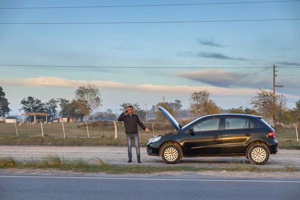Genç Latin bir adam arabasının önünde cep telefonuyla konuşuyor. Yolun kenarında bozulmuş..