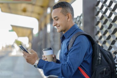 Genç latin adam elinde kahve, elinde kağıt bardakla cep telefonuna bakıyor..