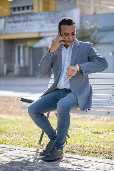 Mladý Latinský Muž Sedí Hranaté Lavičce Mluví Mobilním Telefonu Dívá — Stock fotografie