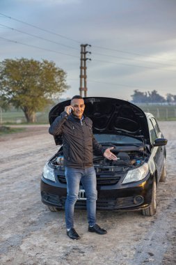 Genç Latin bir adam arabasının önünde cep telefonuyla konuşuyor. Yolun kenarında bozulmuş..