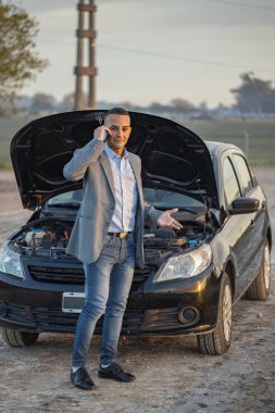 Takım elbiseli genç Latin bir adam yol kenarında arabasının önünde cep telefonuyla konuşuyor..