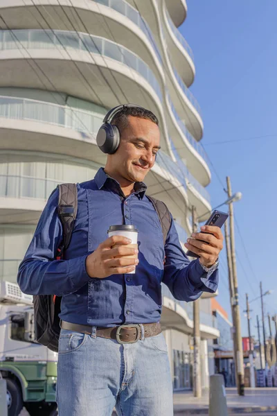 Kulaklıklı genç bir adam cep telefonuna bakıyor ve elinde kahve olan bir kağıt bardak tutuyor..