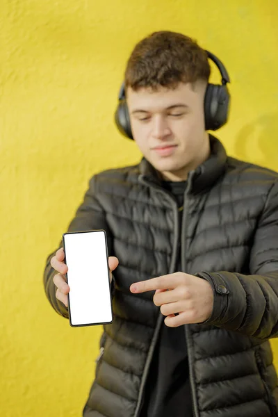 白种人男孩的耳机走焦了 显示出他的手机在黄色背景下被隔离的空白屏幕 — 图库照片