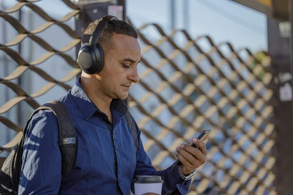 Arkadan görülen otobüs durağındaki cep telefonuna bakan kulaklıklı genç bir Latin adamın orta boy fotoğrafı..