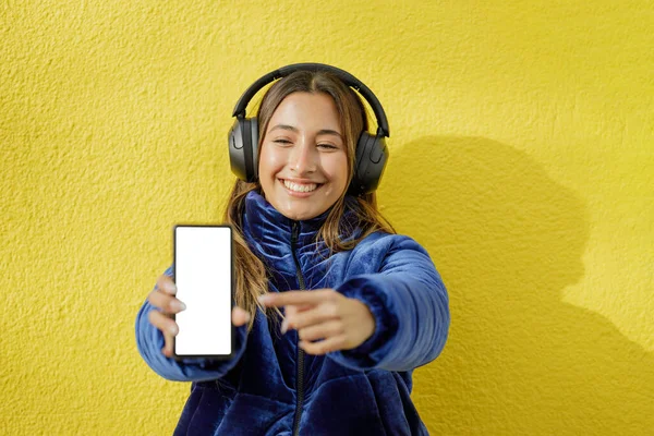 ヘッドフォンを持つラテン語の少女は コピースペースと黄色の背景に隔離された彼女の携帯電話の空白の画面を示しています — ストック写真