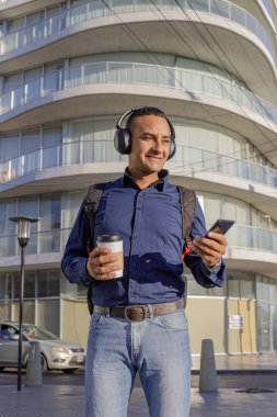 Gülümseyen, kulaklıklı, cep telefonuna bakan ve elinde kahve olan bir kağıt bardak tutan genç Latin bir adam..