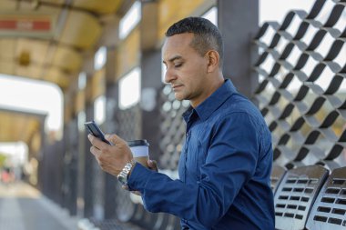 Otobüs durağında elinde kahveyle cep telefonuna bakan genç Latin bir adam..