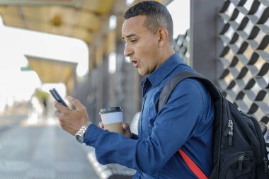 Genç Latin adam cep telefonuna şaşırmış bir suratla bakıyor elinde kahve olan bir kağıt bardak..