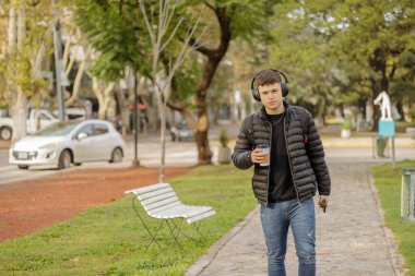 Kulaklıklı ve tek kullanımlık kahvesi olan bir çocuk halka açık bir parkta fotokopi aletiyle yürüyor..