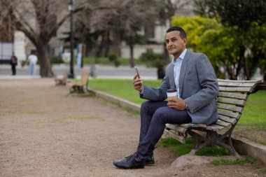 Takım elbiseli bir Latin adam ve tek kullanımlık bir fincan kahve halka açık bir parkta bankta oturuyor..