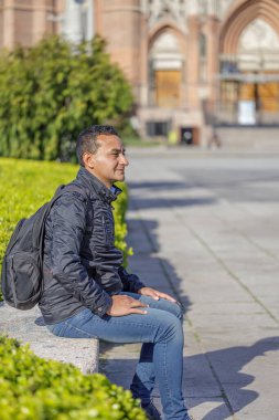 Sırt çantalı genç Latin bir adam halka açık bir parkta bankta oturuyor..