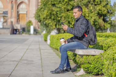Genç Latin bir adam halka açık bir parkta bankta oturuyor cep telefonunu kullanıyor ve kahve içiyor..
