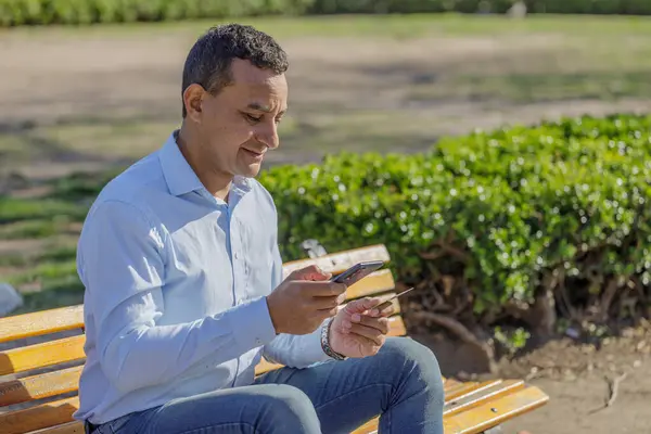 Young Latino man making a purchase with a credit card on his mobile phone in a public park.