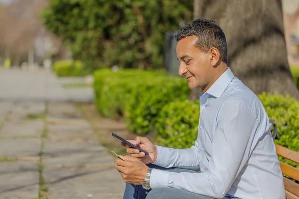 Young Latino man making a purchase with a credit card on his mobile phone sitting on a bench in a public park.
